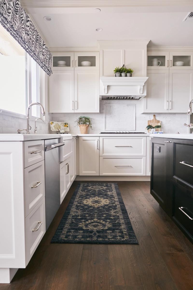 A bright, traditional-style kitchen featuring white inset cabinetry with nickel hardware, a custom range hood with corbels, dark hardwood floors, a navy runner rug, and a farmhouse sink beneath a window with a blue and white Roman shade.