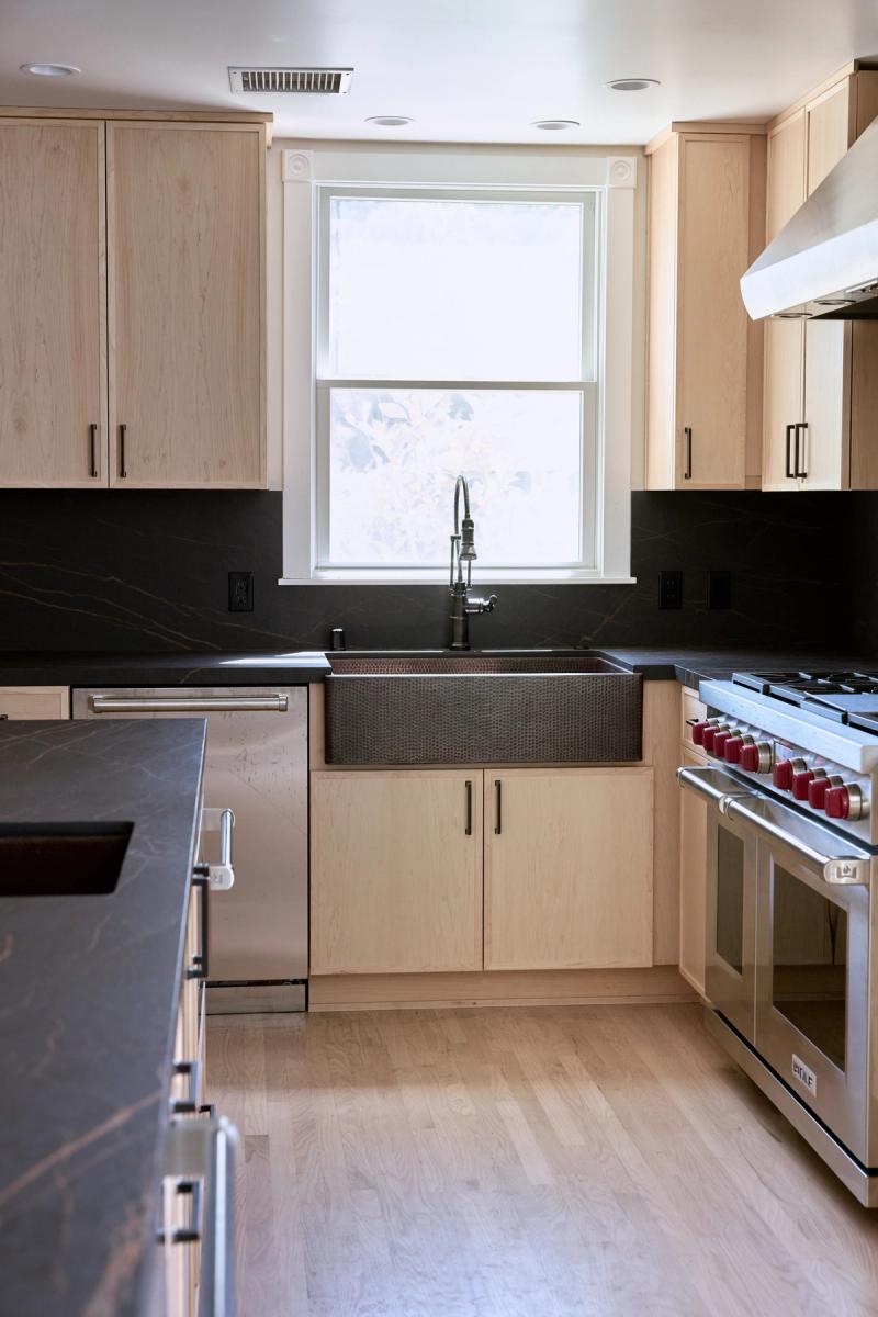 A modern kitchen featuring light white oak cabinetry, dark soapstone countertops and backsplash, a hammered copper farmhouse sink beneath a window, and a professional Wolf range on the right.