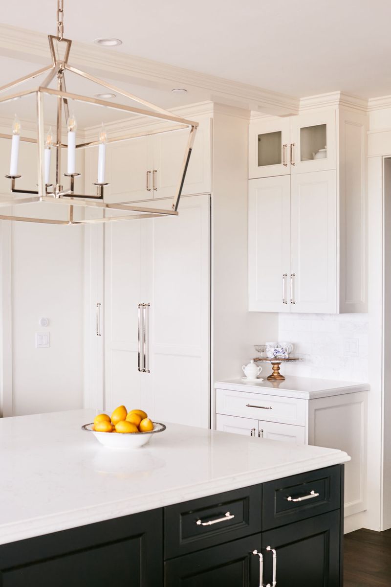 A bright, elegant kitchen featuring white shaker-style wall cabinets and a dark navy or black island with white quartz countertops, polished nickel hardware, a lantern-style chandelier, and a bowl of lemons on the island.