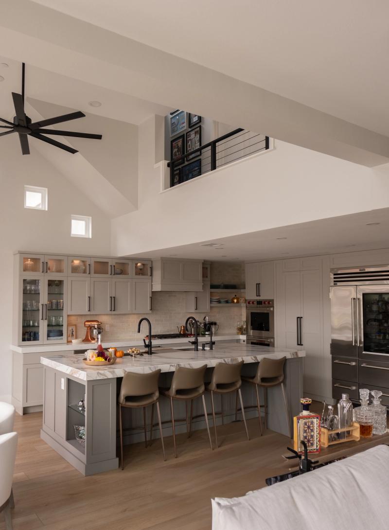 A spacious open-concept kitchen featuring gray shaker cabinets, a large marble-top island with bar seating, high vaulted ceilings with a ceiling fan, and a second-floor loft visible above.