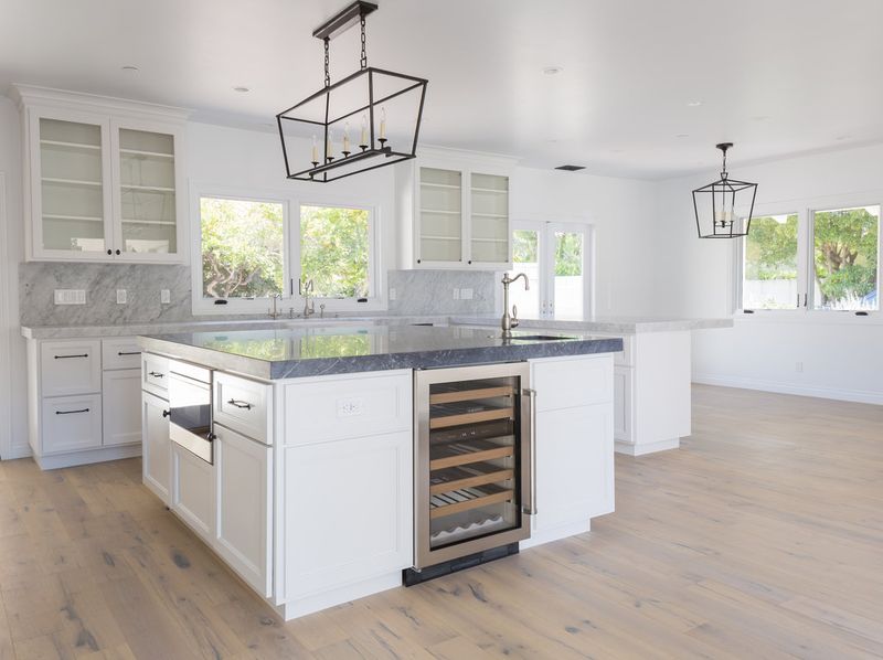 A bright, spacious white shaker-style kitchen featuring a large center island with dark marble countertops, a built-in wine cooler, glass-front upper cabinets, and black cage pendant lighting over light hardwood floors.