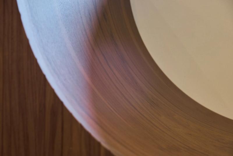 An extreme close-up of a circular or oval cutout in a wood panel, showing fine wood grain texture with warm brown tones and soft light reflections.