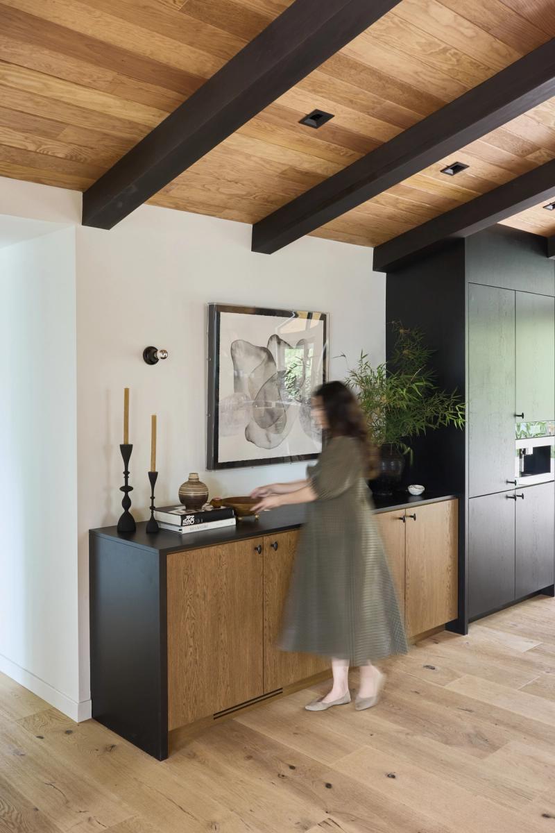 A woman in a green dress walks past a custom oak and matte black sideboard cabinet in a modern home with exposed wood beam ceiling and dark cabinetry in the background.