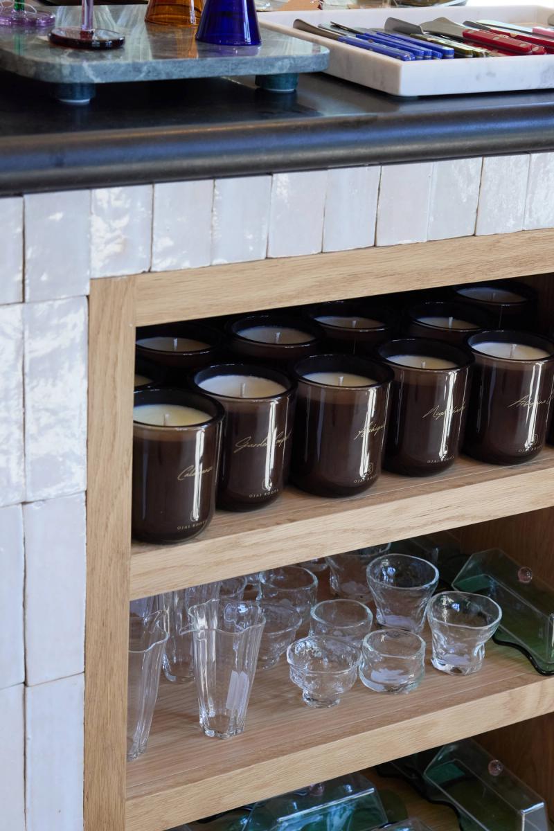 A light oak open shelving unit with white zellige tile surround, displaying a collection of dark amber glass candles labeled 'Ojai Roots' on the upper shelf and clear glassware on the lower shelf.