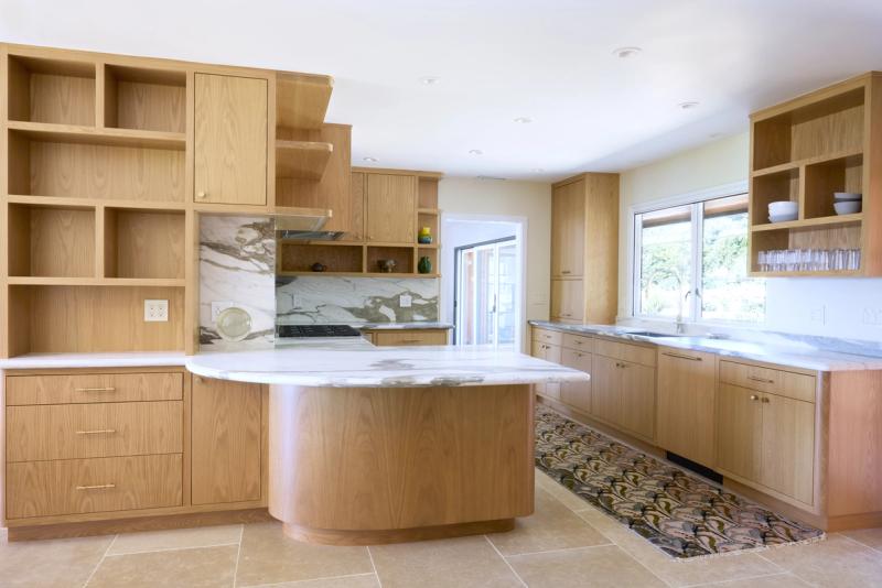 A bright, open kitchen featuring warm white oak shaker cabinets, black marble slab backsplash, stainless steel Sub-Zero refrigerator, and a professional range, set beneath a vaulted white ceiling with hardwood floors.