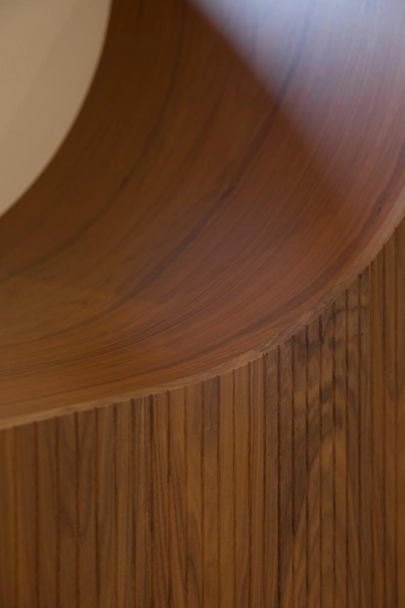 A close-up detail shot of a curved walnut wood surface transitioning into vertical fluted wood paneling, showcasing custom craftsmanship and rich wood grain.