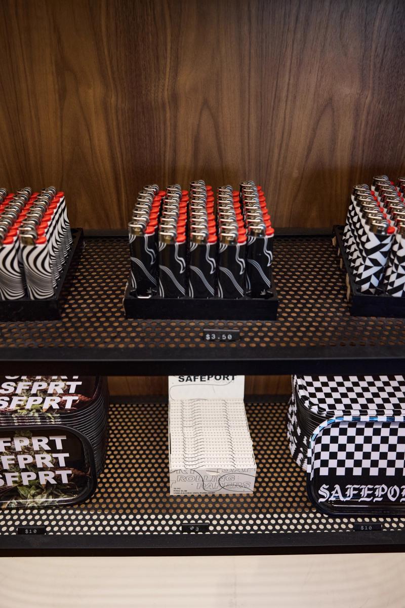 Close-up of a retail display unit featuring black perforated metal shelves and a warm walnut wood back panel, stocked with lighters and rolling paper accessories.
