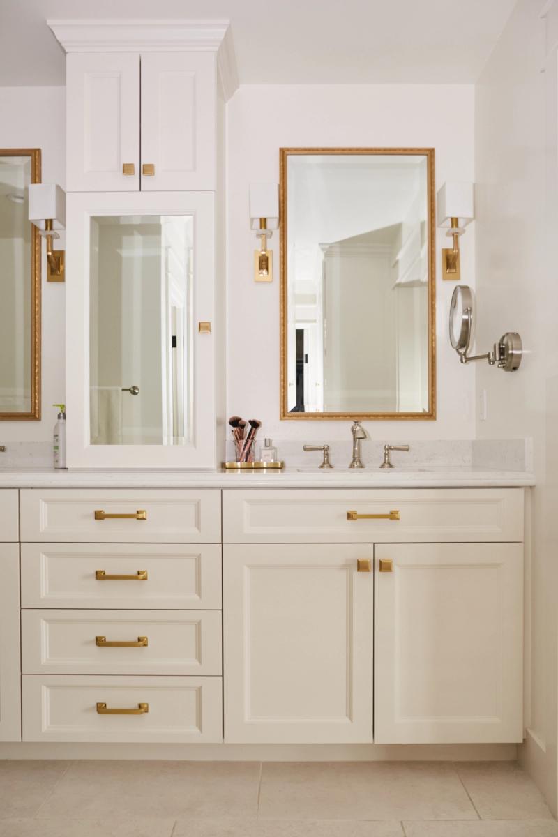 A bright, elegant bathroom vanity featuring white raised-panel cabinetry with brushed gold hardware, white marble countertop, gold-framed mirrors, and wall sconces. The vanity includes a tower cabinet, four drawers, and lower cabinet doors.