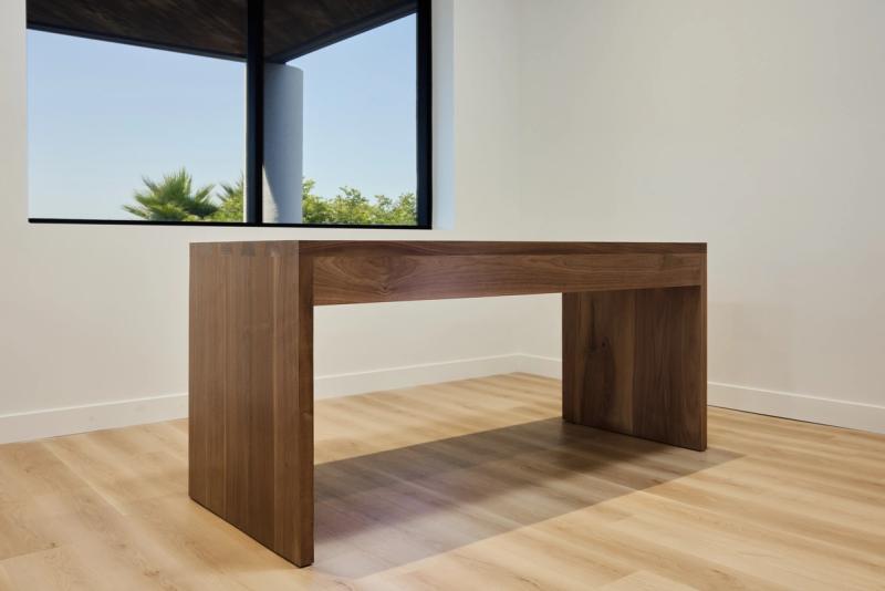 A sleek walnut waterfall-edge desk with panel sides sits in a bright, minimalist room with light wood floors and a large window overlooking greenery.