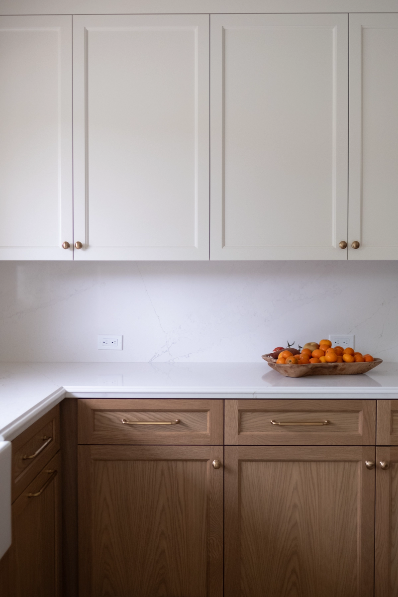 A clean, modern kitchen featuring white shaker upper cabinets with brass knobs and natural white oak lower cabinets with brass bar pulls, topped with a white quartz countertop and matching marble-look backsplash. A wooden bowl of oranges sits on the counter.