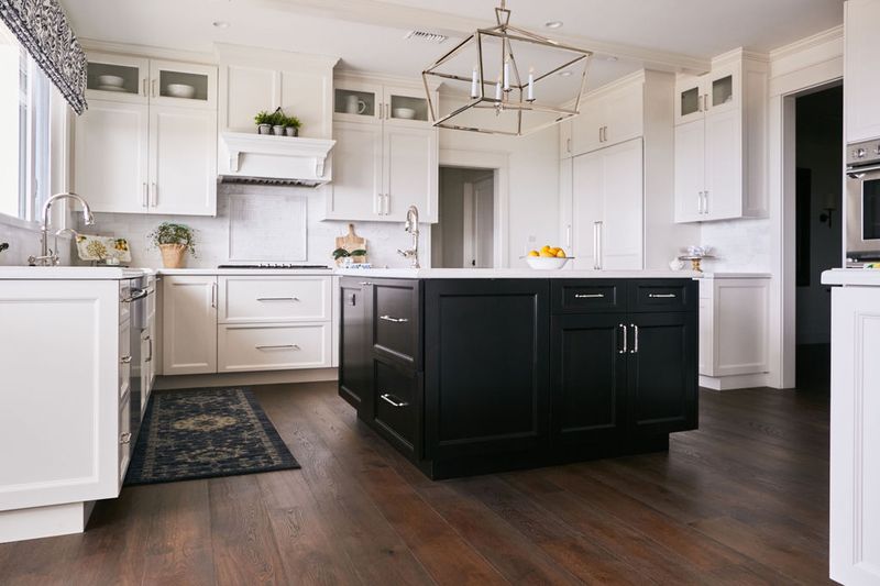 A spacious custom kitchen featuring white shaker-style perimeter cabinets with a contrasting dark black kitchen island, white quartz countertops, hardwood floors, and a modern chrome lantern pendant light.