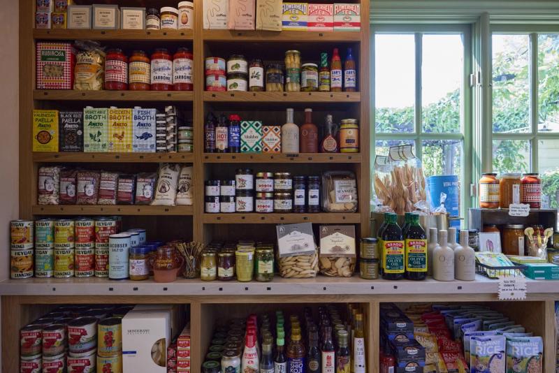 A well-organized specialty grocery or pantry display featuring floor-to-ceiling custom oak shelving units filled with artisan food products, jars, and pantry goods, with a large counter below holding additional items and a window with garden views in the background.