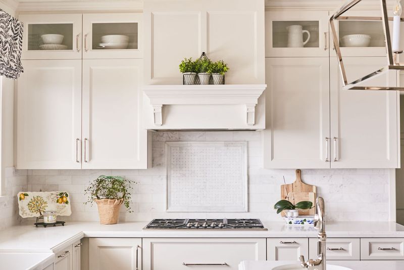 A bright, elegant white kitchen featuring custom inset cabinetry with glass uppers, a decorative wood range hood surround, marble tile backsplash, and quartz countertops accented with fresh greenery and silver hardware.