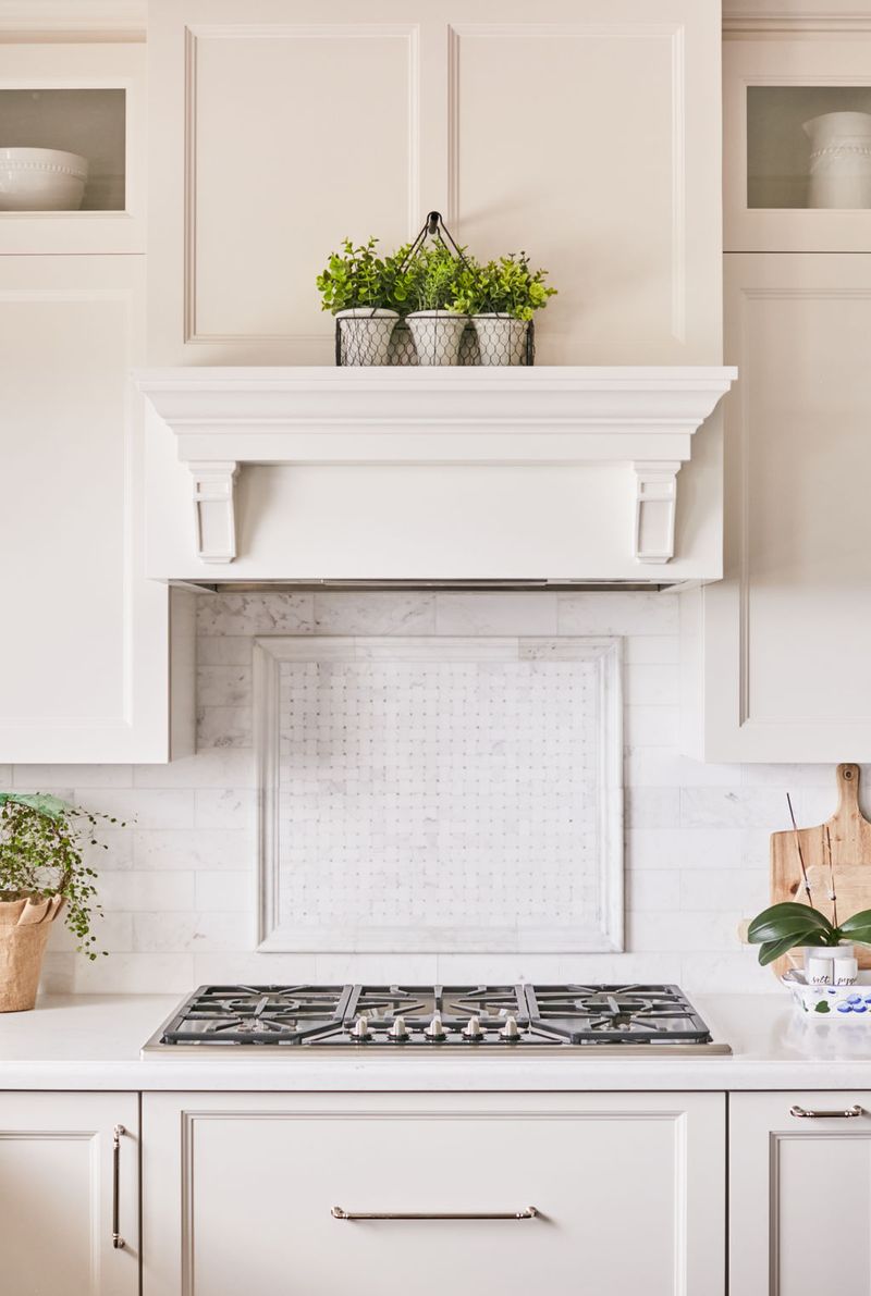 A bright, elegant kitchen featuring a custom white painted range hood with corbel details and mantel shelf, marble tile backsplash with basketweave inset, and cream shaker-style cabinetry with brushed nickel hardware.