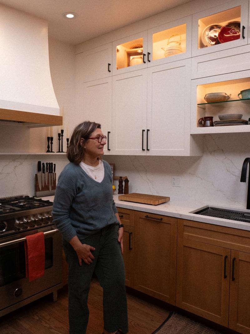 A woman stands in a beautifully remodeled kitchen featuring white upper cabinets with glass fronts and interior lighting, natural wood lower cabinets, a marble slab backsplash, and a professional stainless steel range with a plaster hood.