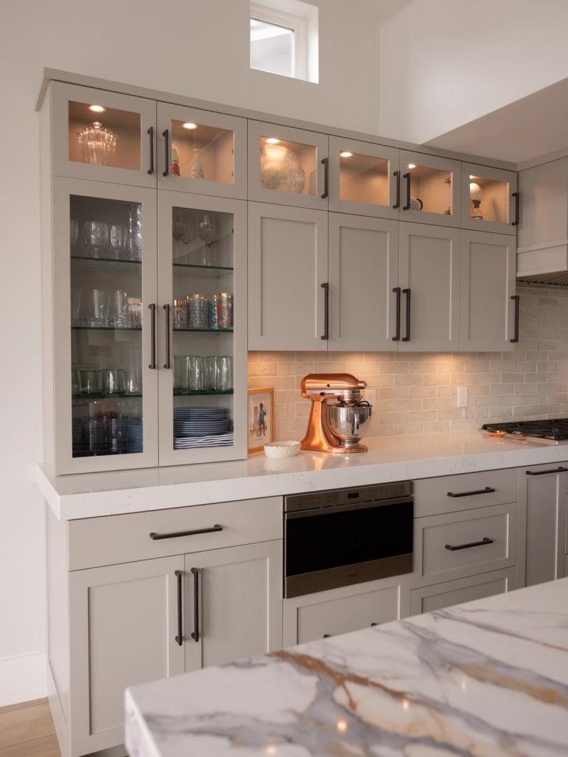 A beautifully designed kitchen featuring floor-to-ceiling gray shaker cabinets with glass-front doors displaying glassware, dark bronze hardware, white quartz countertops, a subway tile backsplash, and a copper KitchenAid stand mixer on the counter.