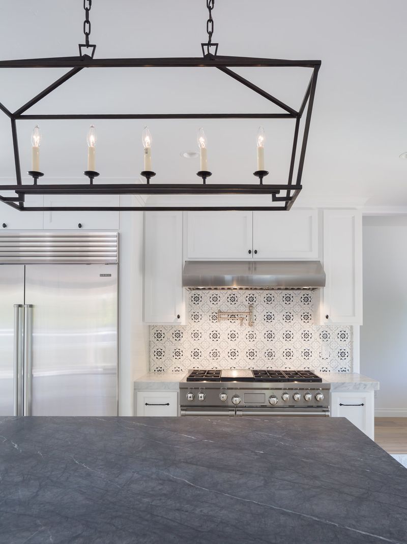 A bright, elegant kitchen featuring white shaker cabinets, a stainless steel professional range with decorative patterned tile backsplash, a built-in refrigerator, dark soapstone island countertop, and a black iron linear chandelier overhead.