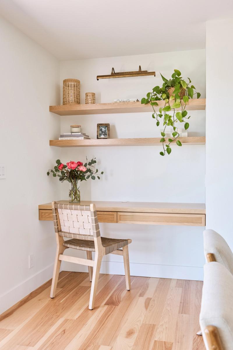 A minimalist home office nook featuring a floating white oak desk with drawers, two matching wall-mounted shelves styled with plants, books, and woven baskets, and a woven leather chair.