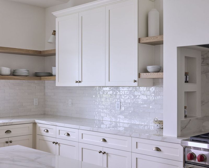 A bright, all-white custom kitchen featuring shaker-style cabinets with brushed nickel hardware, open natural wood shelving, white marble countertops, and a glossy subway tile backsplash. A stainless steel range is visible on the right side.