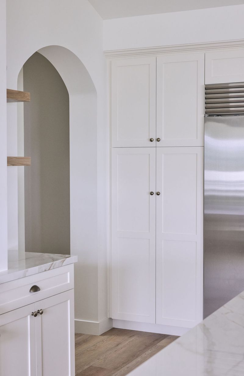 A bright white kitchen featuring tall floor-to-ceiling inset pantry cabinets with antique brass knobs, an arched built-in niche with open wood shelving, and a stainless steel refrigerator. The space has light hardwood floors and marble countertops.