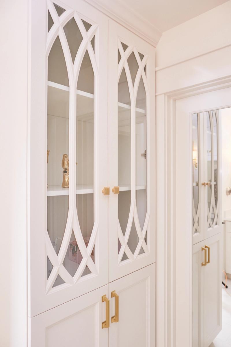 Elegant white built-in cabinetry with gothic arch lattice glass doors and brushed brass hardware, featuring open display shelves with gold decorative accents.