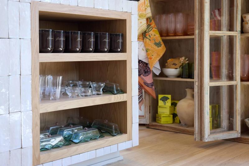 A light oak built-in shelf unit recessed into a white handmade tile wall, displaying glassware and serving pieces in a retail or showroom setting. Adjacent vintage cabinet and colorful kitchen textiles are visible in the background.