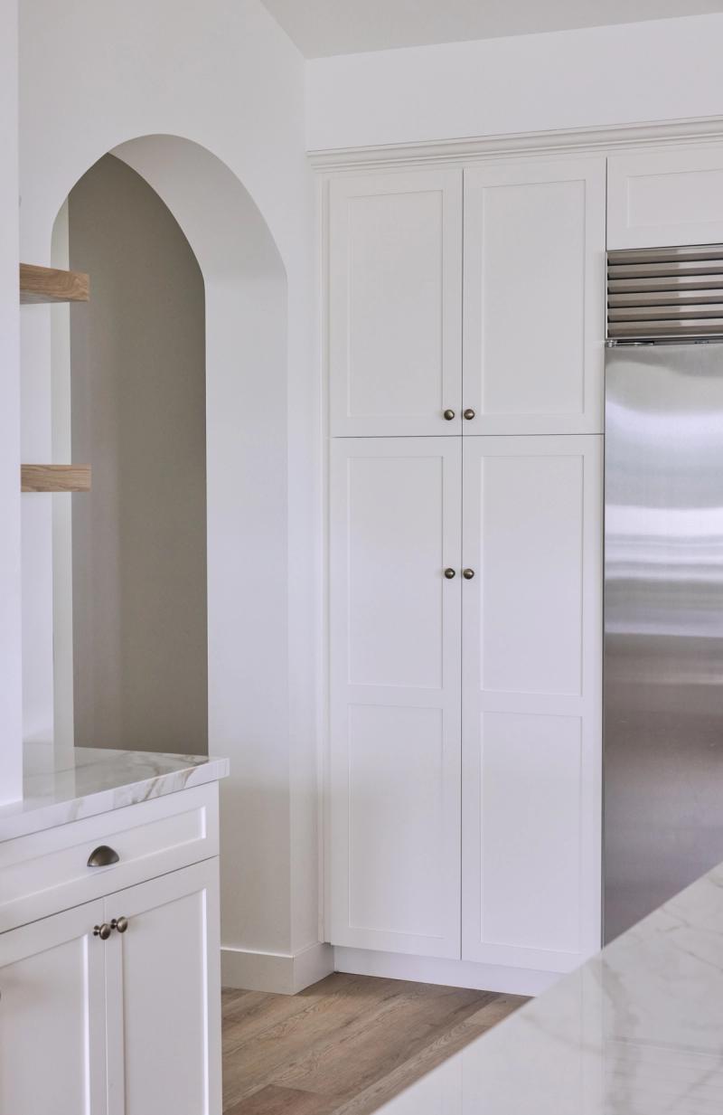 A bright white kitchen featuring tall floor-to-ceiling inset pantry cabinets with antique brass knobs, an arched built-in niche with open wood shelving, and a stainless steel refrigerator. The space has light hardwood floors and marble countertops.