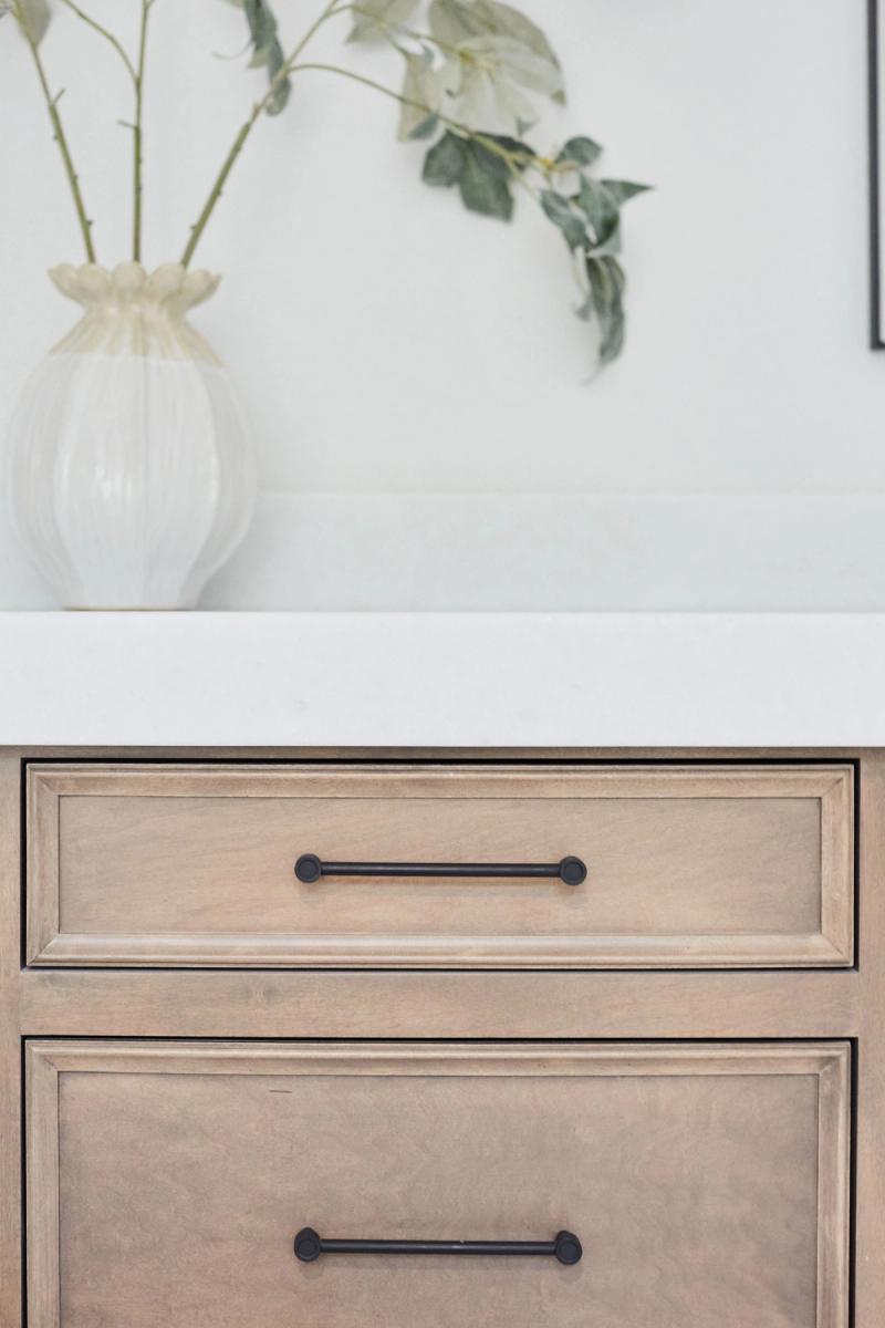 Close-up of a light wood bathroom vanity with two drawers featuring matte black bar pulls, topped with a white quartz countertop and a ribbed white ceramic vase with greenery.