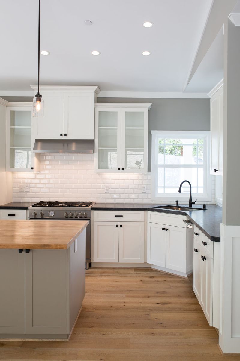 A bright, transitional kitchen featuring white shaker cabinets, dark soapstone countertops, a gray island with butcher block top, white subway tile backsplash, and light oak hardwood floors.