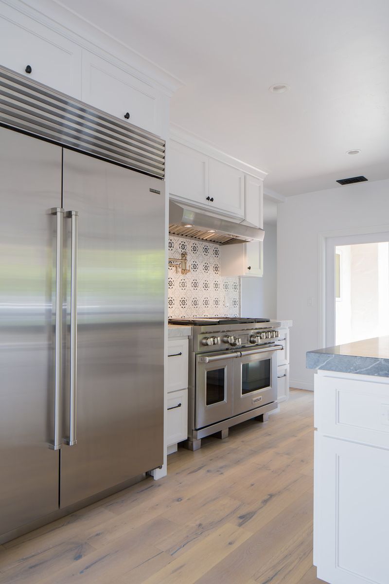 A bright, modern kitchen featuring white shaker-style cabinetry, a stainless steel Sub-Zero refrigerator, a Wolf dual-fuel range with decorative tile backsplash, and light hardwood flooring.