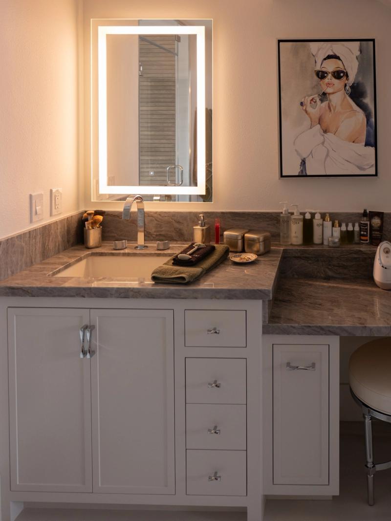 A elegantly designed bathroom vanity featuring white shaker-style cabinets with chrome hardware, gray marble countertop, an illuminated LED mirror, and an integrated makeup vanity area with stool. Beauty products and accessories are neatly arranged on the countertop.
