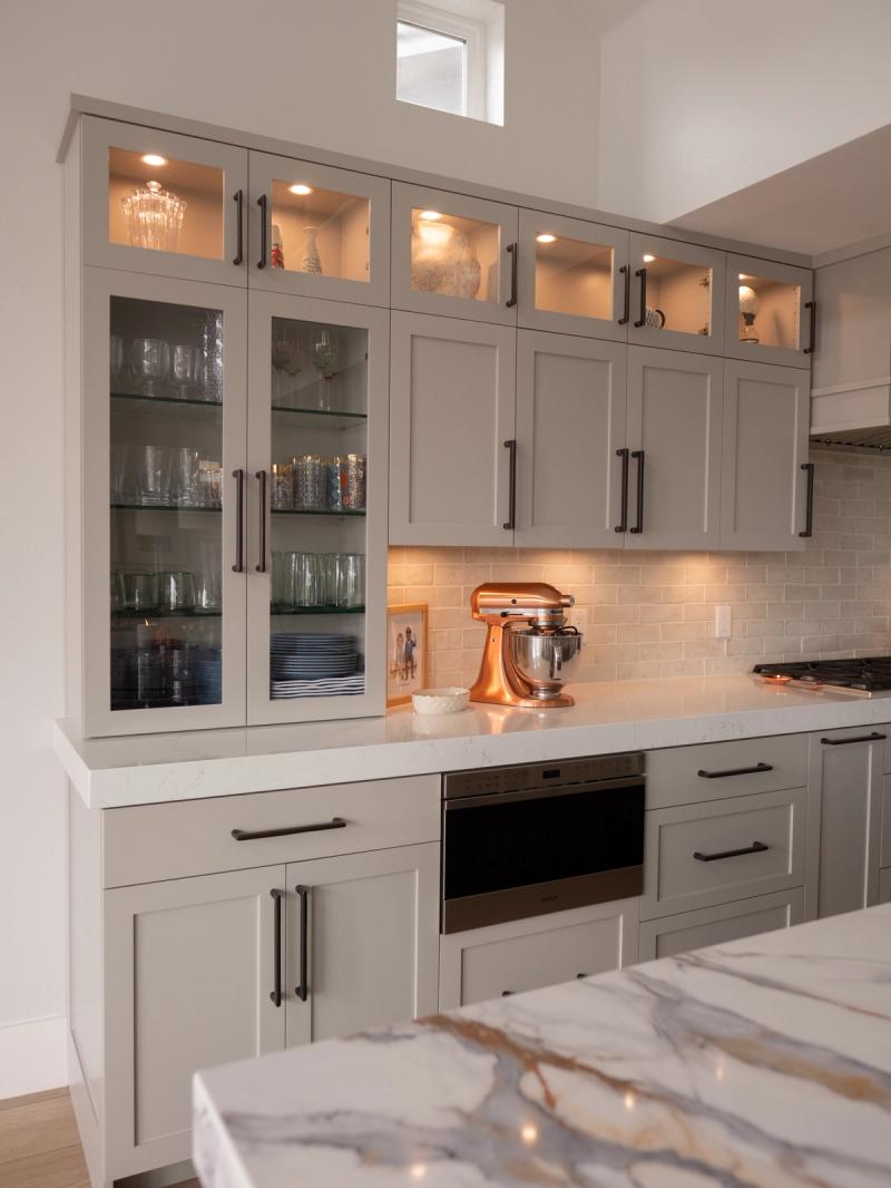 A beautifully designed kitchen featuring floor-to-ceiling gray shaker cabinets with glass-front doors displaying glassware, dark bronze hardware, white quartz countertops, a subway tile backsplash, and a copper KitchenAid stand mixer on the counter.