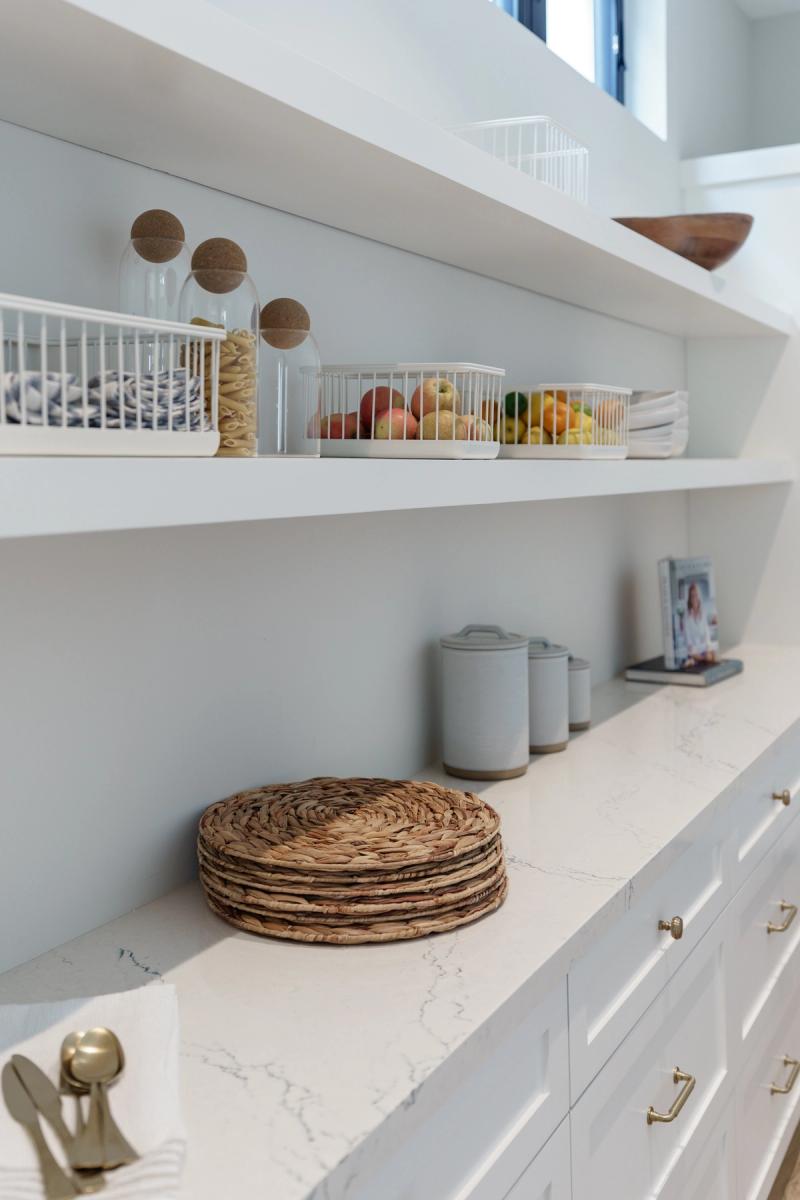 A bright, organized pantry with white open shelving holding wire baskets of fruit, glass canisters, and ceramic containers, above a marble-topped cabinet with white shaker drawers and brass hardware.