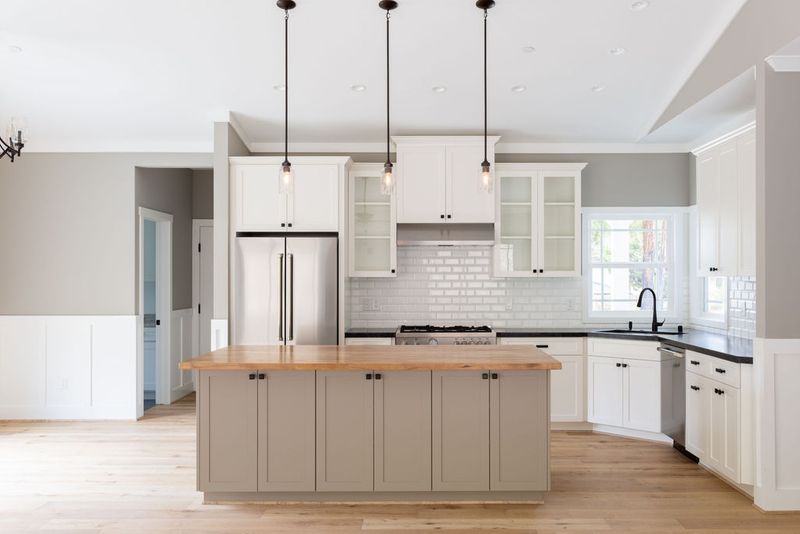 A bright, open kitchen featuring white upper cabinets, greige shaker-style island with butcher block countertop, subway tile backsplash, stainless steel appliances, and three pendant lights over the island.