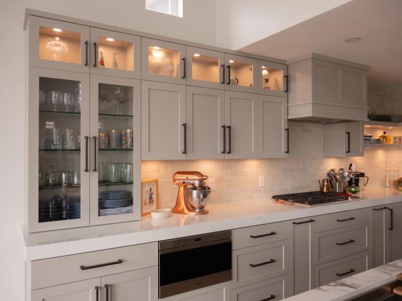 A beautifully designed kitchen featuring floor-to-ceiling gray shaker cabinets with dark hardware, illuminated glass-front upper cabinets displaying glassware, white quartz countertops, a subway tile backsplash, and a copper KitchenAid stand mixer on the counter.