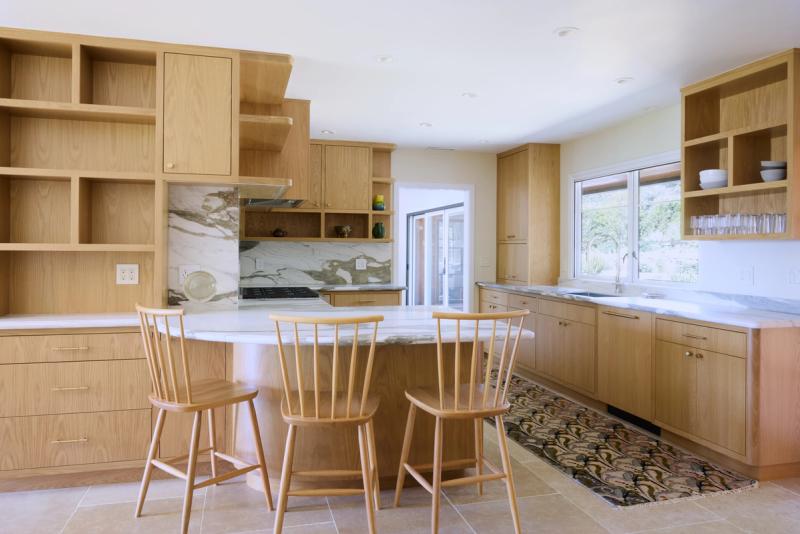 A bright, modern kitchen featuring custom natural oak cabinetry with open shelving, a curved marble-topped island with three Windsor bar stools, a marble backsplash, and a large window overlooking greenery.