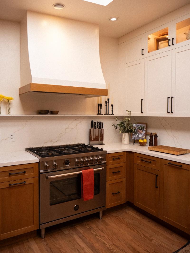 A custom kitchen featuring warm wood lower cabinets, white upper cabinets with black hardware, a professional gas range, and a large plaster range hood with wood trim accents.