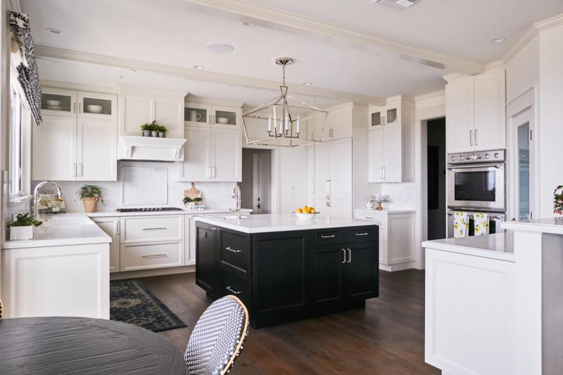 A bright, spacious kitchen featuring white shaker cabinets with chrome hardware, a dark navy-black center island with white quartz countertop, hardwood floors, and a geometric chrome pendant light overhead.