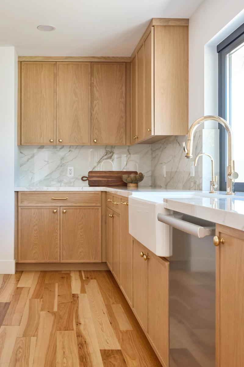 A bright kitchen featuring light white oak shaker cabinets with brass hardware, a white marble slab backsplash, white quartz countertops, a farmhouse sink with a gold faucet, and warm wood flooring.