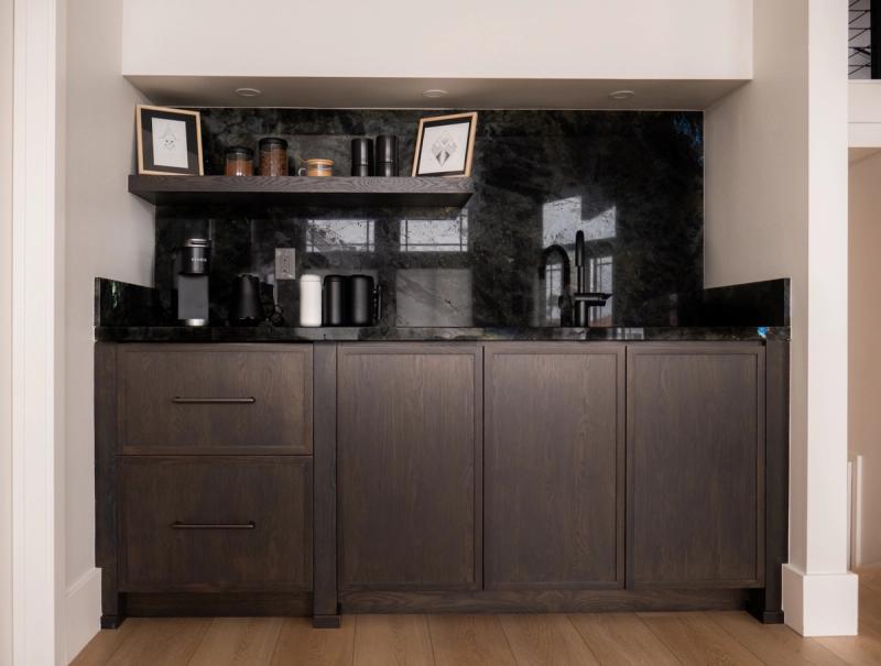A built-in coffee bar station featuring dark stained oak cabinetry with a dramatic black stone backsplash and countertop, open floating shelf with accessories, and matte black fixtures including a Keurig coffee maker and sink faucet.