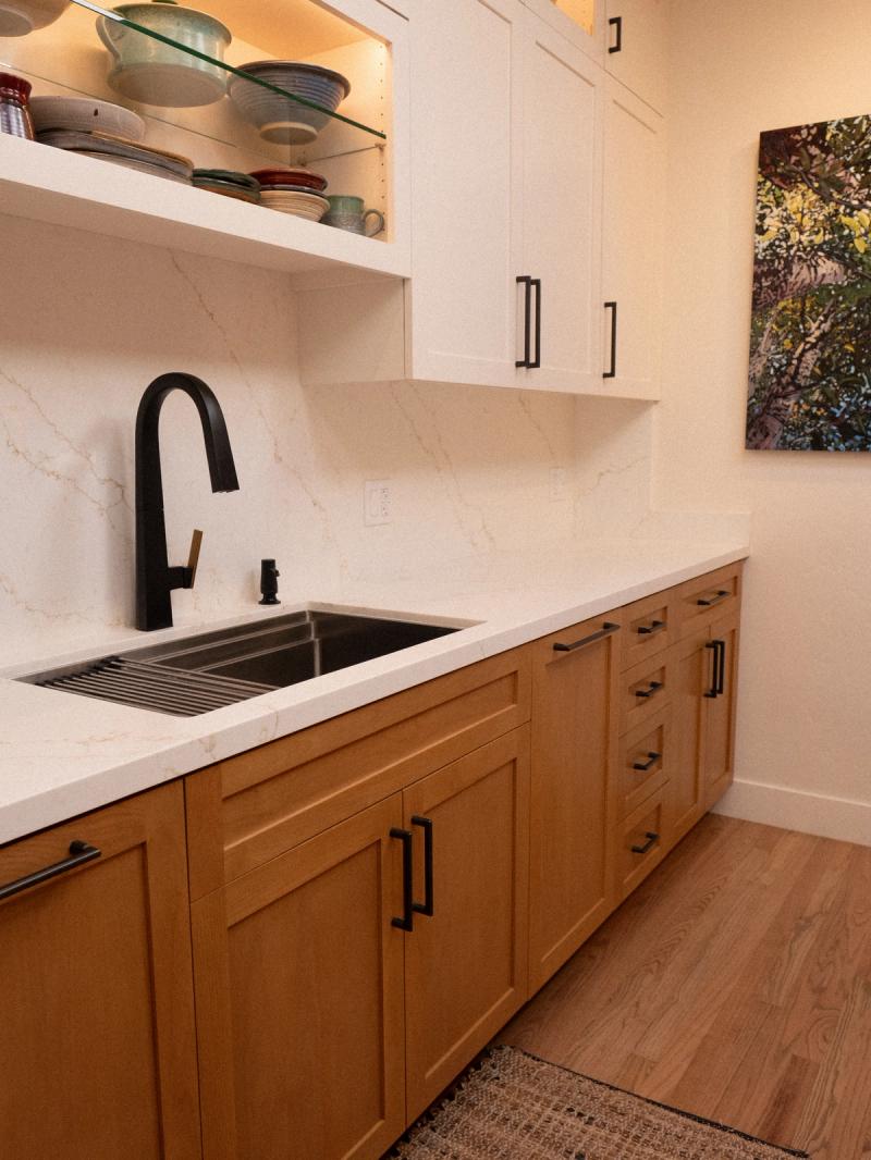 A modern kitchen featuring warm honey-toned lower cabinets and white upper cabinets with black hardware, a white quartz countertop and backsplash, and a matte black pull-down faucet over an undermount sink.