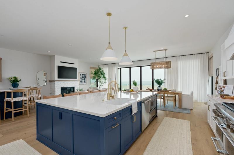 A bright, open-concept coastal kitchen featuring a large navy blue island with white quartz countertops, brass hardware and faucet, pendant lighting, and floor-to-ceiling windows with an ocean view beyond the dining and living area.