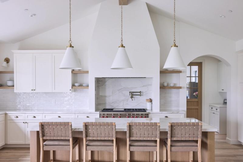 A bright, airy custom kitchen featuring white inset cabinetry, open oak shelving, a dramatic arched plaster range hood with marble slab backsplash, a large island with woven leather bar stools, and three white cone pendant lights with brass hardware.