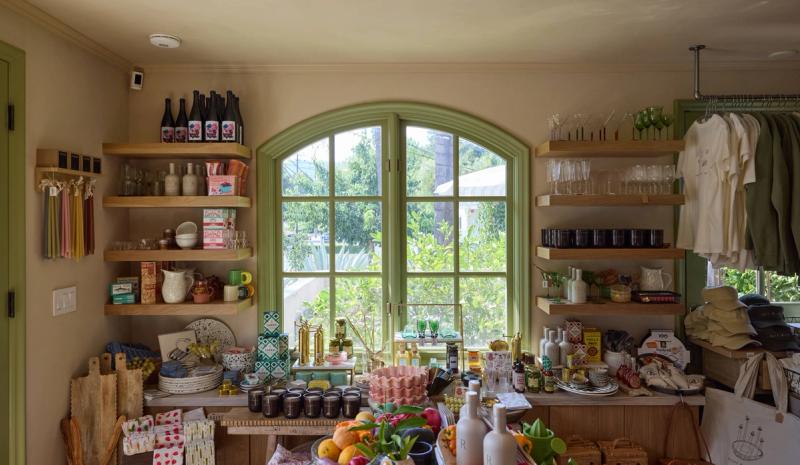 A charming retail shop interior featuring open wood floating shelves displaying bottles, glassware, candles, and pantry goods flanking a green-painted arched window. A central display table is filled with curated products including ceramics, fresh fruit, and specialty items.