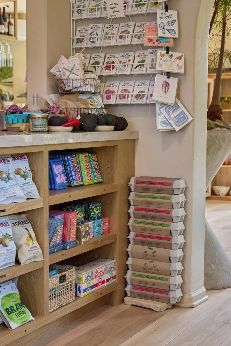 A light oak shelving unit built into a retail store corner, displaying snacks, teas, and specialty goods, with seed packets and greeting cards on a wall rack above the counter.