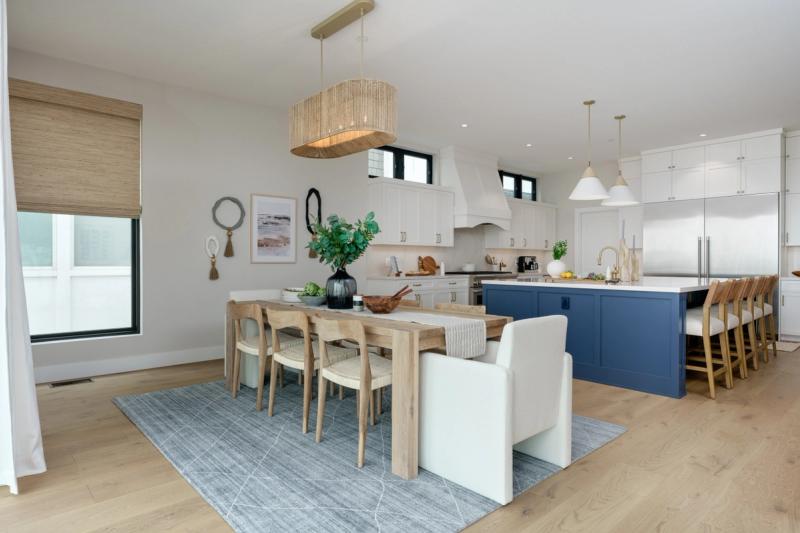 An open-concept kitchen and dining area featuring white shaker cabinetry, a large navy blue island with bar seating, a natural wood dining table, and a woven pendant light above the dining space.
