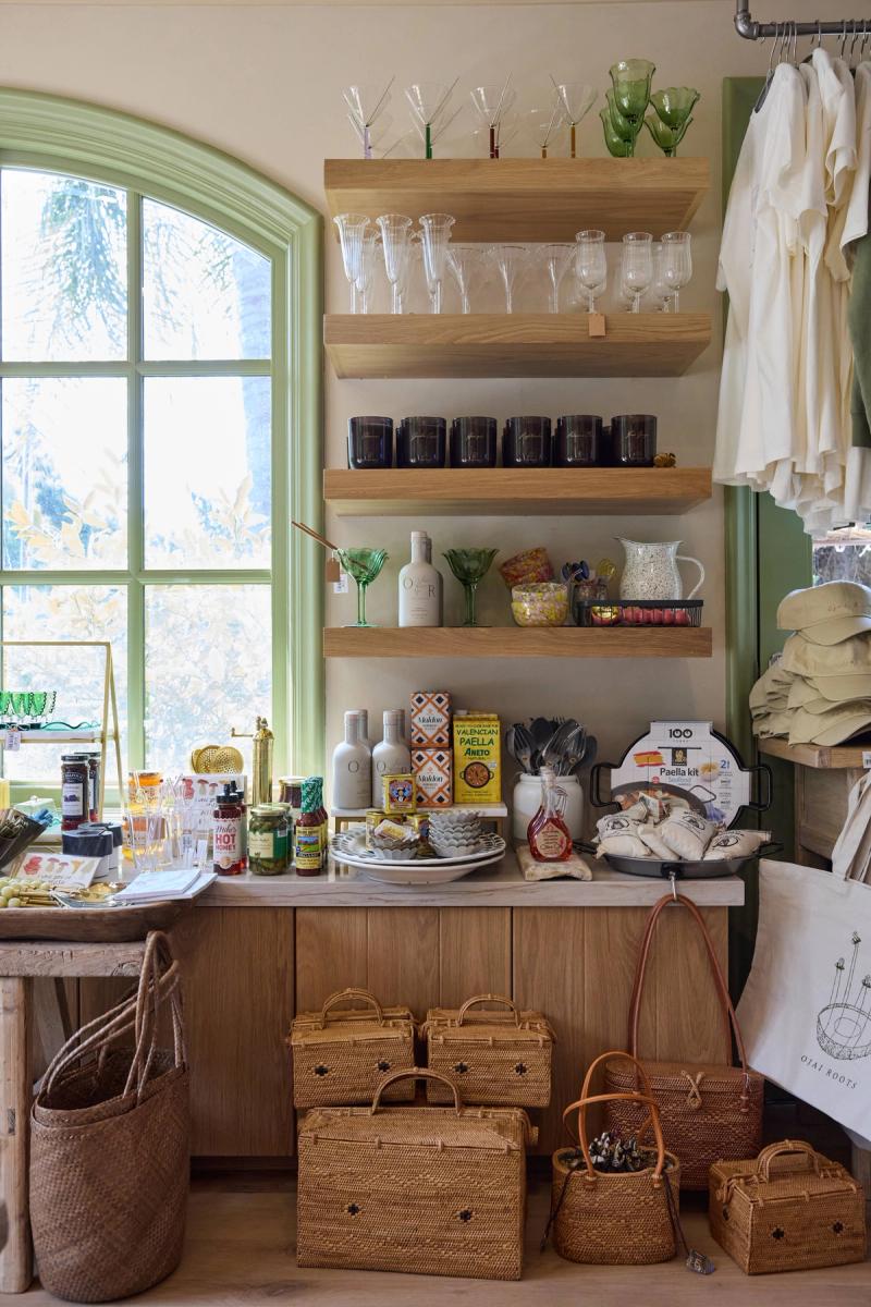 A charming retail shop interior featuring custom oak floating shelves mounted on a green-trimmed wall, displaying glassware, candles, and pantry goods, with wicker baskets arranged below a marble-topped wooden counter beside an arched window.