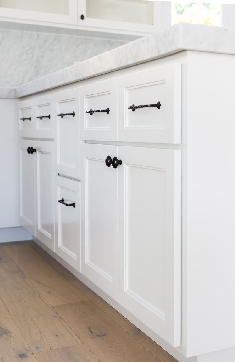 White shaker-style bathroom vanity cabinets with marble countertop and matte black twisted-bar pulls and knobs, set against light hardwood flooring.