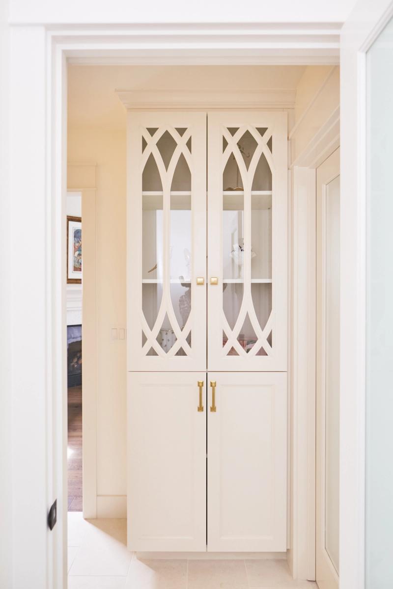A tall cream-painted built-in cabinet with decorative Gothic arch glass upper doors and solid lower doors, accented with brushed brass hardware, set into a hallway nook.