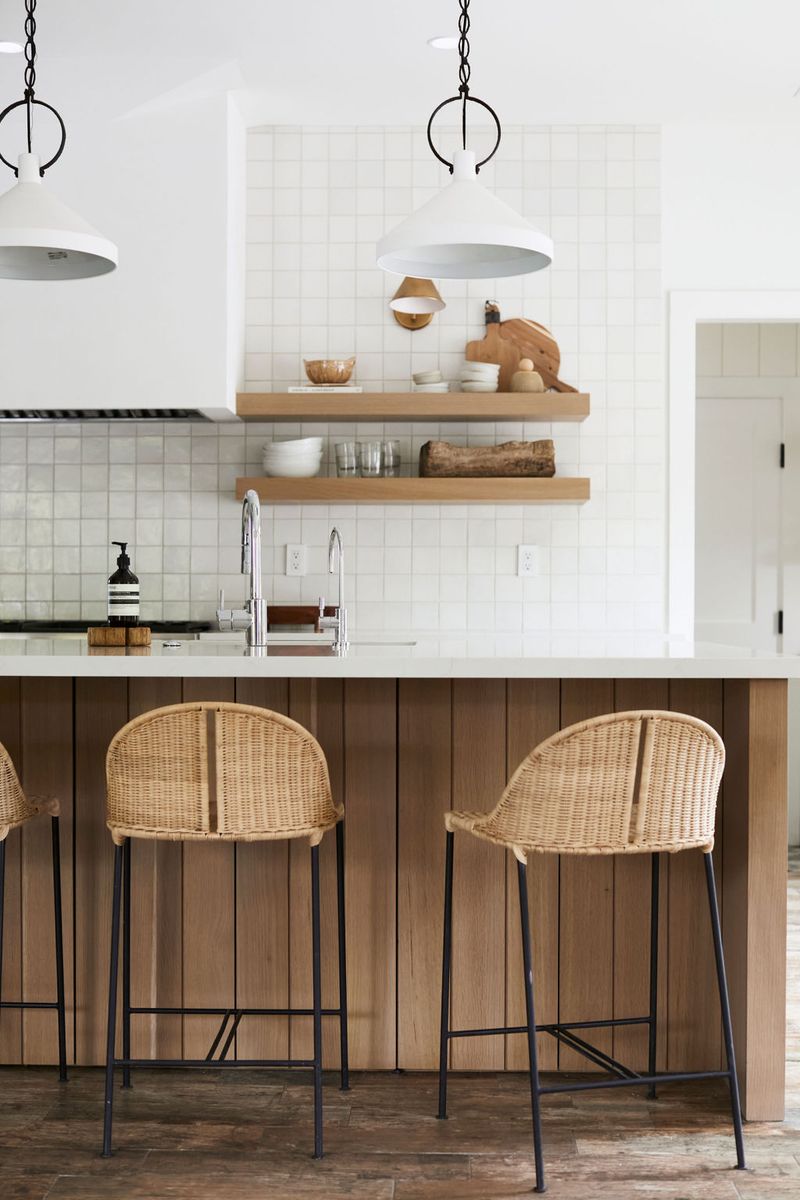 A bright, modern kitchen featuring a wood-paneled island with rattan bar stools, open oak floating shelves, square white tile backsplash, and white pendant lights.